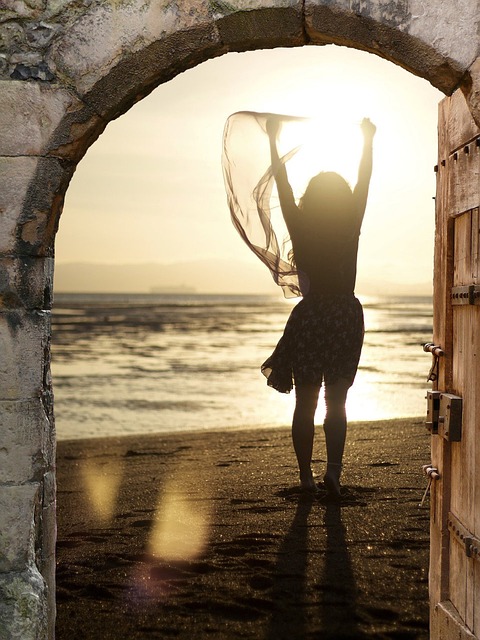 Frau Silhouette am Strand. Zu sehen ist ein Sonnenuntergang am Strand und ein Frauen-Silhouette, die die Hände hebt und dabei flatter ein leichtes durchsichtiges Tuch. Man sieht die Frau durch einen Torbogen