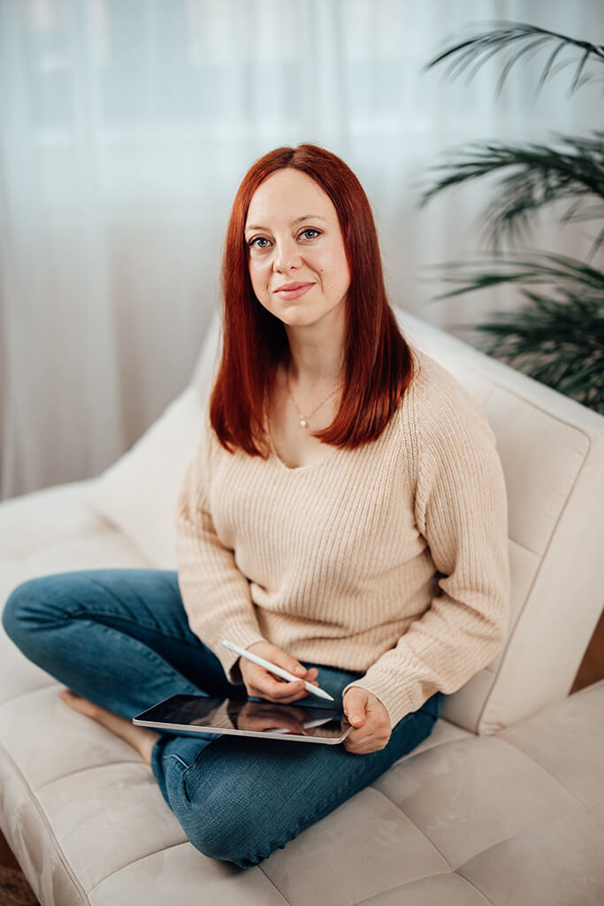 Profilbild Vanessa Hofer-Kauschinger, schmunzelnd, im Schneidersitz sitzend auf einer cremefarbenen Couch, Ipad und Stift in der Hand, gekleidet mit einer dunkelblauen Jeans und einem beigen Pullover