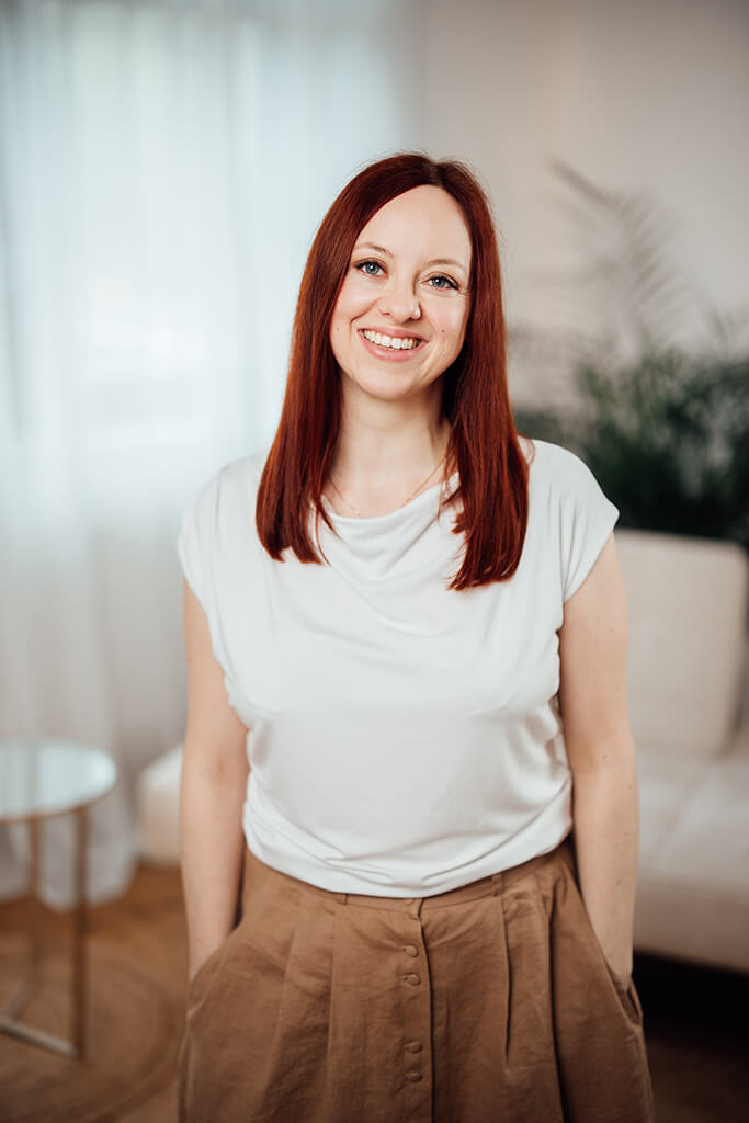 Portrait von Vanessa Hofer-Kauschinger, lächelnd, bekleidet mit einem braunen Rock und einem weißem T-Shirt, stehend im Hintergrund ist eine cremefarbene Coach zu sehen und Pflanzen