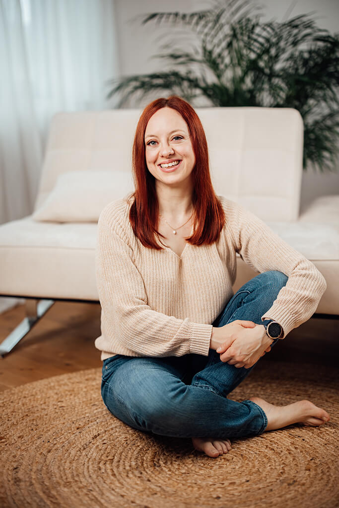 Portrait von Vanessa Hofer-Kauschinger, lächelnd, mit kupferfarbenen langen Haaren, sitzend auf einem runden Sisal-Teppich und angelehnt an eine cremefarbene Couch, barfuß, mit einer dunkelblauen Jeans und eine beigen Strickpullover bekleidet.